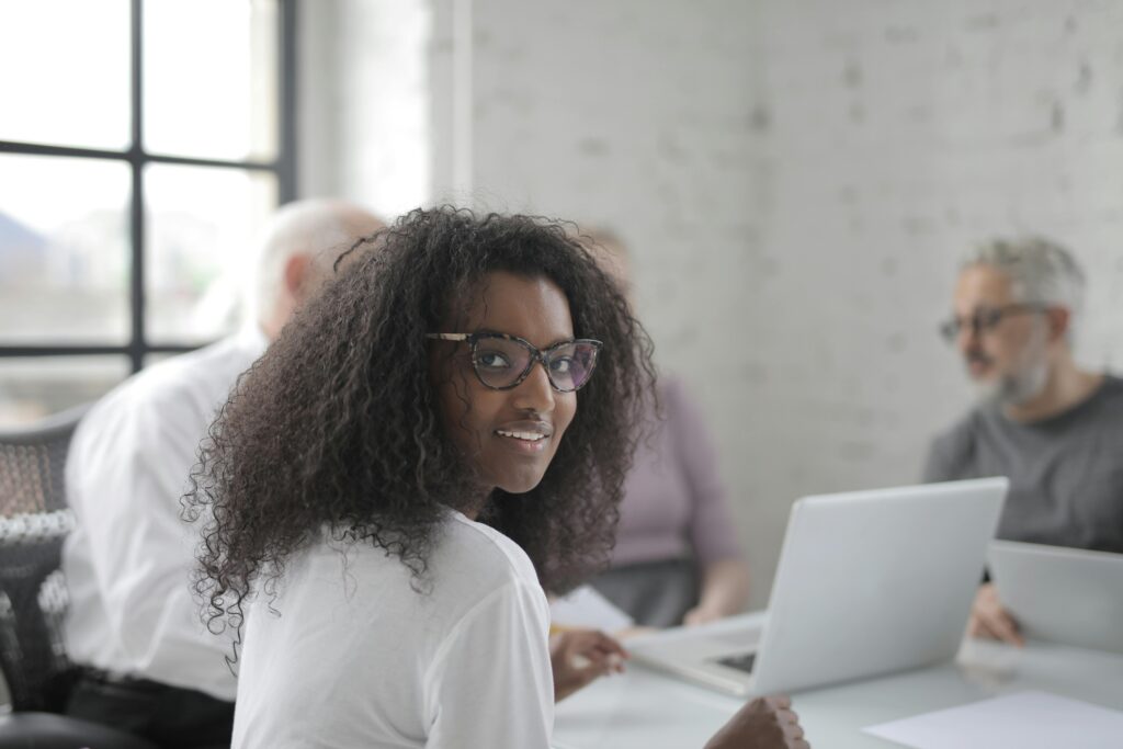 Positive ethnic woman wearing formal clothes and eyeglasses smiling while working with colleagues in modern workspace and looking at camera